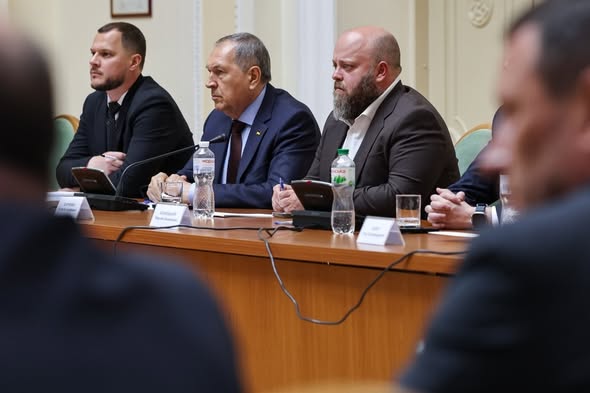Три посадовці в ділових костюмах сидять за довгим дерев'яним столом президії з мікрофонами та пляшками води під час офіційного засідання, а на передньому плані видно розмиті силуети інших учасників заходу