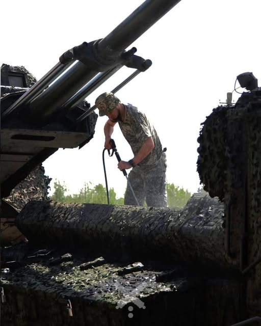 Військовослужбовець у камуфляжній формі та панамі миє артилерійську установку водою зі шланга високого тиску просто неба на тлі дерев