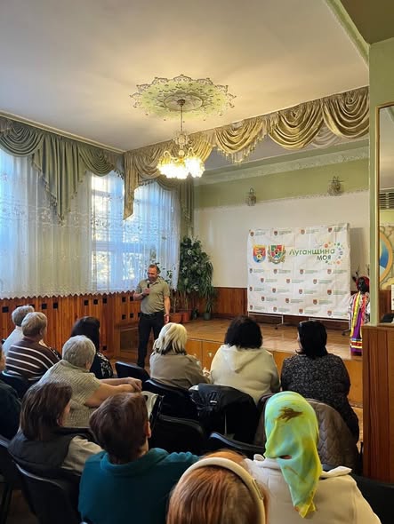 Чоловік із мікрофоном виступає на сцені залу перед сидячою авдиторією слухачів, а праворуч від нього встановлено інформаційний стенд із написом «Луганщина моя».