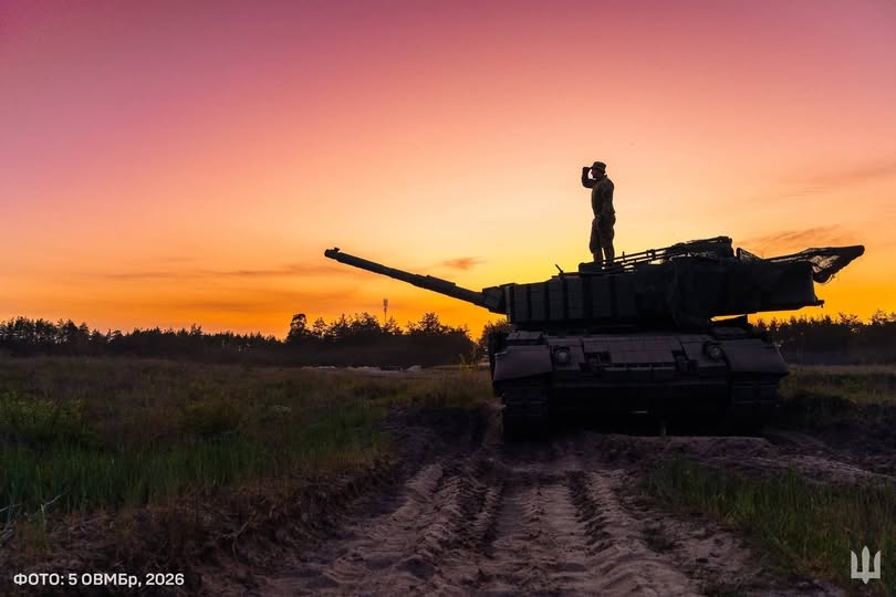 Силует військового танка з бійцем на башті, який дивиться вглиб поля на тлі яскравого помаранчево-рожевого заходу сонця.