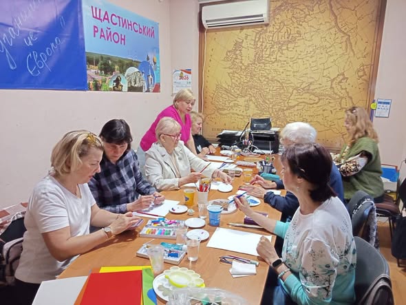Група переважно літніх жінок сидить за довгим столом у кабінеті під час творчого заняття з малювання, де на стінах розміщені плакат «Щастинський район» та велика старовинна мапа
