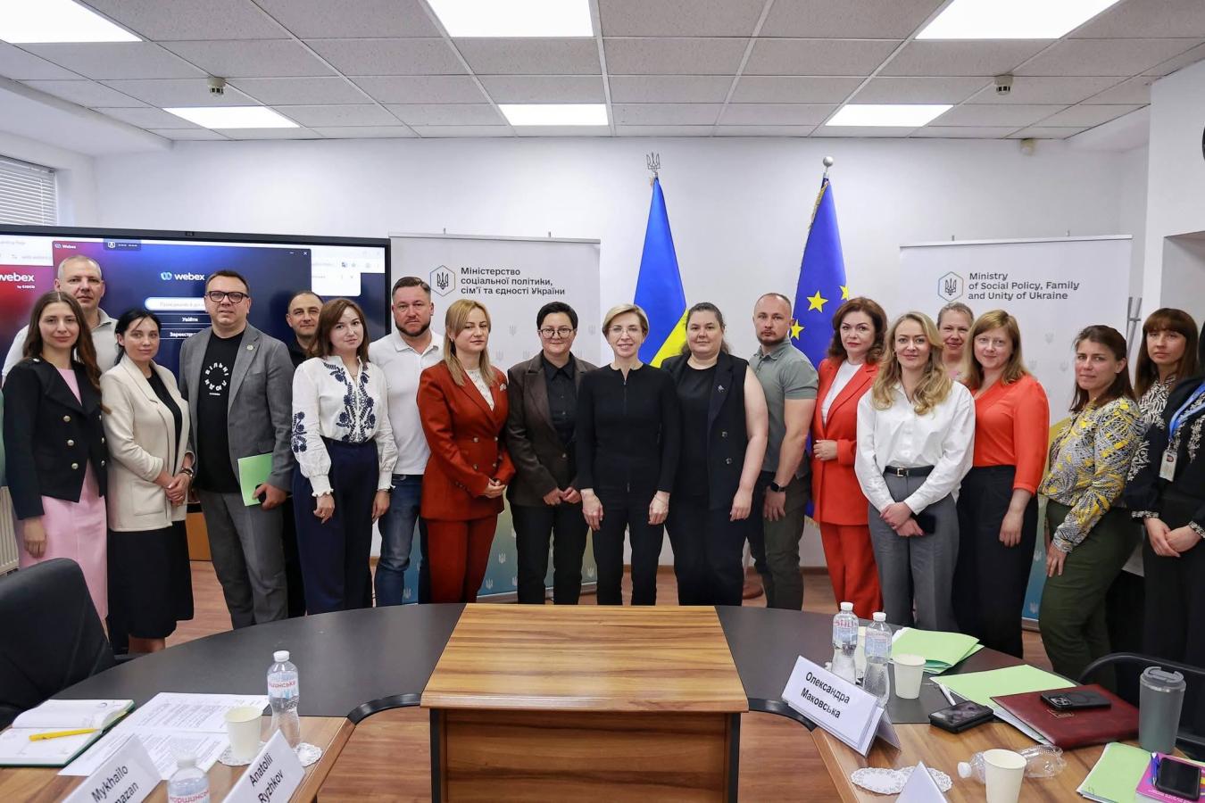 Група людей стоїть у конференційній залі для спільного фото на тлі прапорів України та Європейського Союзу, інформаційних стендів Міністерства соціальної політики та великого екрана