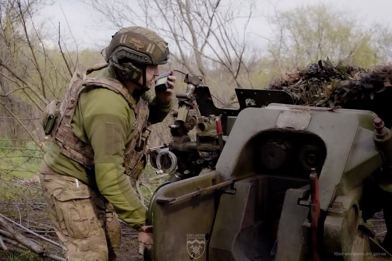 Український військовослужбовець у камуфляжній формі та шоломі з навушниками говорить по рації, перебуваючи на бойовій позиції біля великої артилерійської установки серед дерев