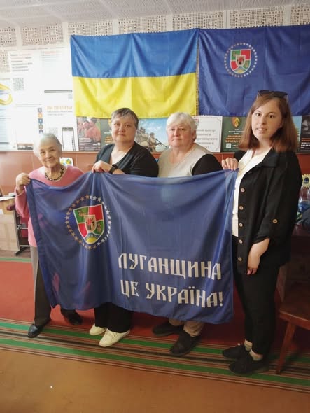 Колаж із чотирьох фотографій демонструє патріотичний захід просто неба, де група людей похилого та молодого віку позирує біля виставкових стендів із фотографіями, прапором Луганщини, виробами ручної роботи та банером із написом «Луганщина — це Україна»
