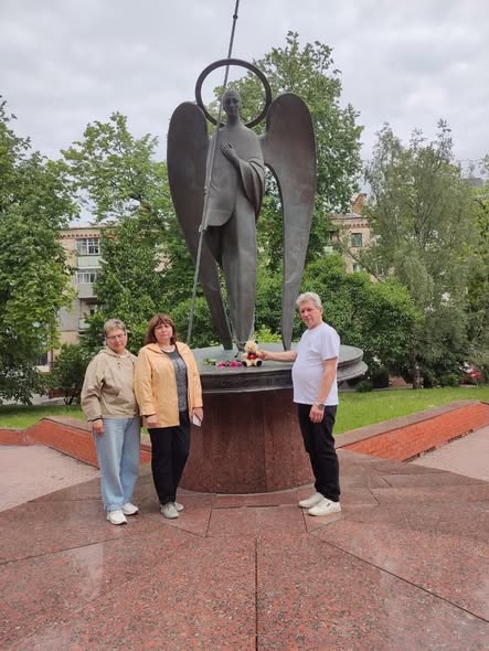 Дві жінки та чоловік стоять біля підніжжя високого бронзового пам'ятника ангелу з німбом і довгим списом, де чоловік кладе м'яку іграшку ведмедика поруч із квітами на знак пам'яті