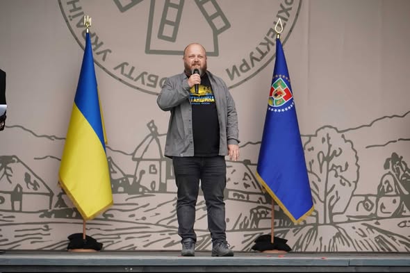 Чоловік із мікрофоном у руках виступає на сцені між державним прапором України та прапором Луганської області на тлі банера з традиційними українськими мотивами