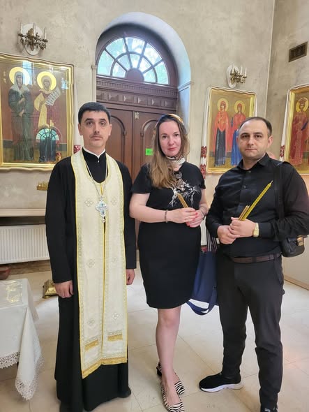 Православний священник у чорному підряснику та світлій єпітрахілі стоїть у храмі поруч із жінкою та чоловіком у темному одязі, які тримають у руках тонкі церковні свічки на тлі ікон та вхідних дверей.