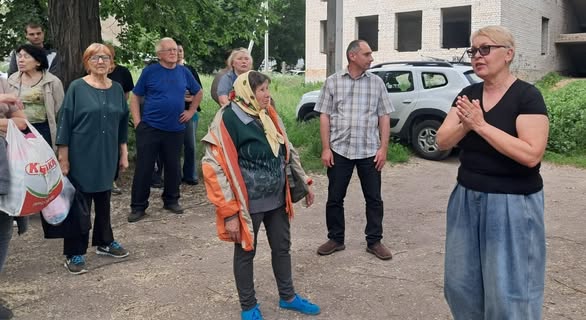 Група людей різного віку стоїть на вулиці біля цегляної будівлі та автомобіля, слухаючи жінку в окулярах та чорній футболці, яка щось емоційно розповідає та тримає руки разом