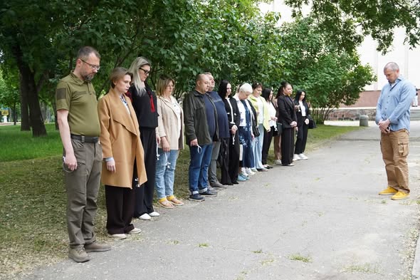 Група людей вишикувалася в шеренгу на асфальтованій доріжці в парку, схиливши голови під час хвилини мовчання, тоді як навпроти них стоїть чоловік у блакитній сорочці та штанах карго