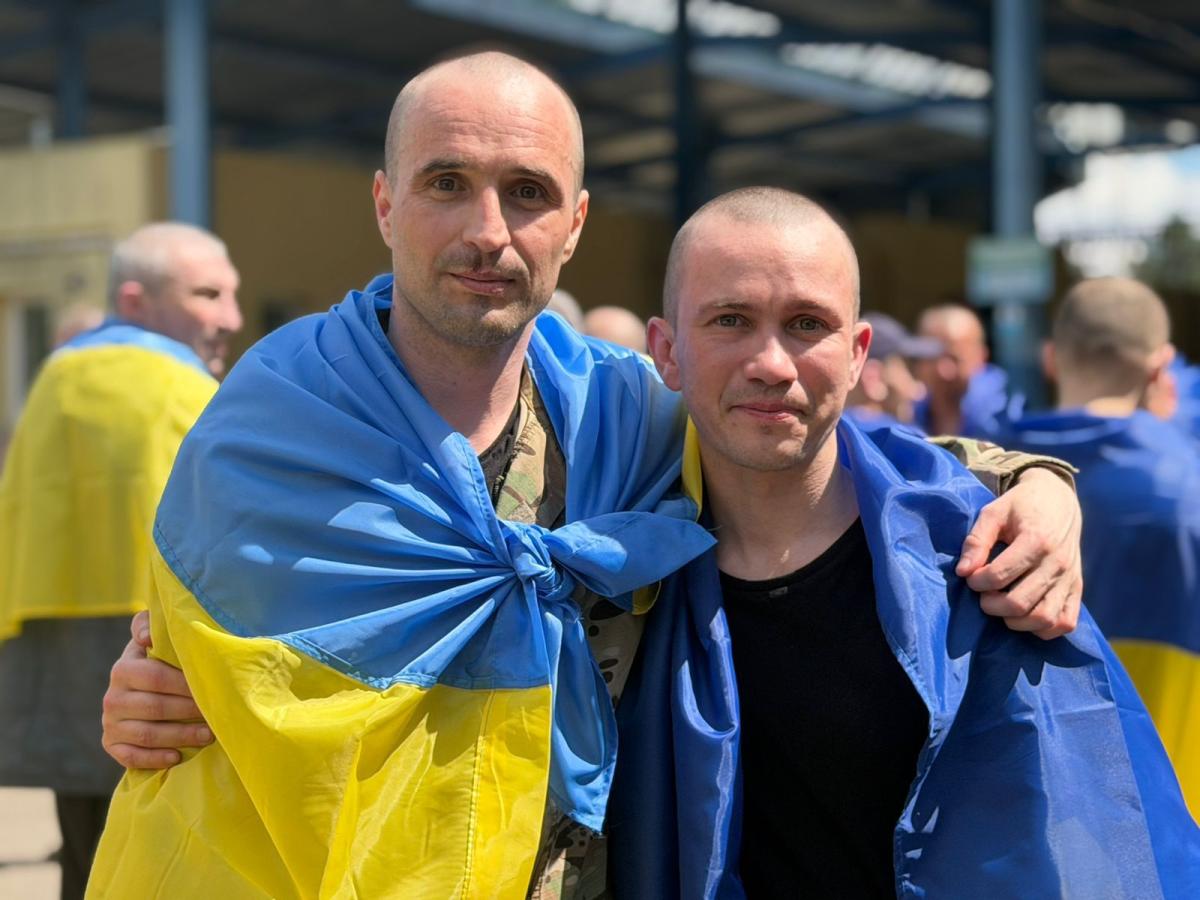 Два звільнених з полону українських військовослужбовці з поголеними головами дружньо обіймаються за плечі на вулиці, загорнувшись у синьо-жовті прапори України