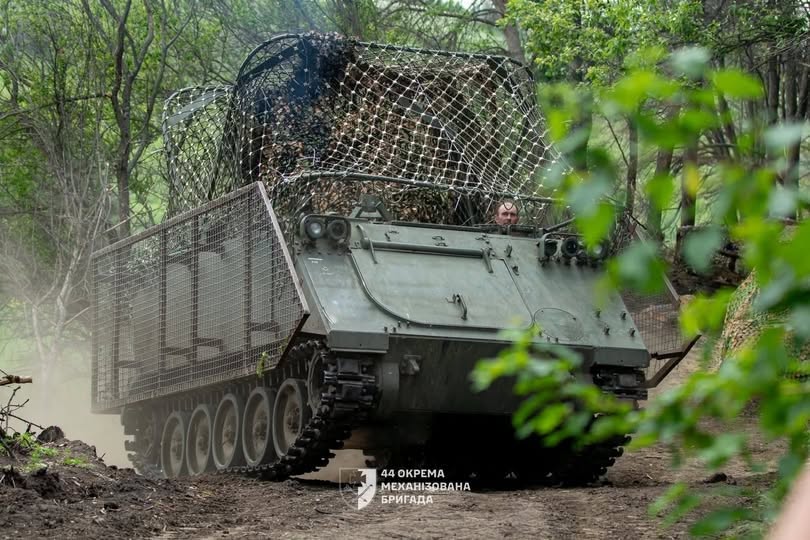 Гусеничний бронетранспортер М113, додатково захищений металевими решітками та маскувальною сіткою «мангал» на даху, рухається ґрунтовою дорогою серед лісового масиву, а з люка визирає водій-механік