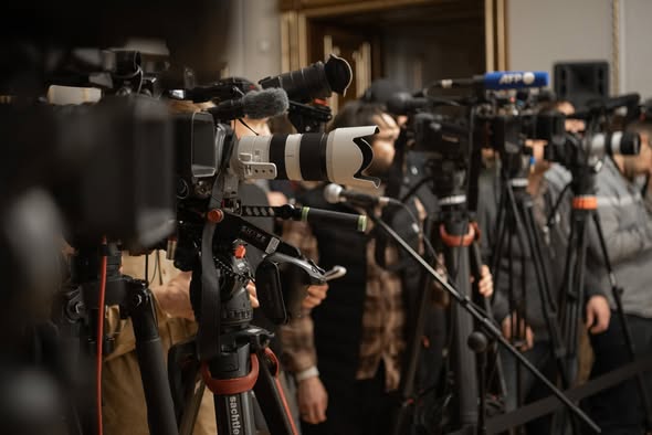 Ряд професійних телевізійних та фотокамер на штативах, обладнаних мікрофонами та великими об'єктивами, встановлено в приміщенні для фіксації медійної події за участю журналістів та операторів.