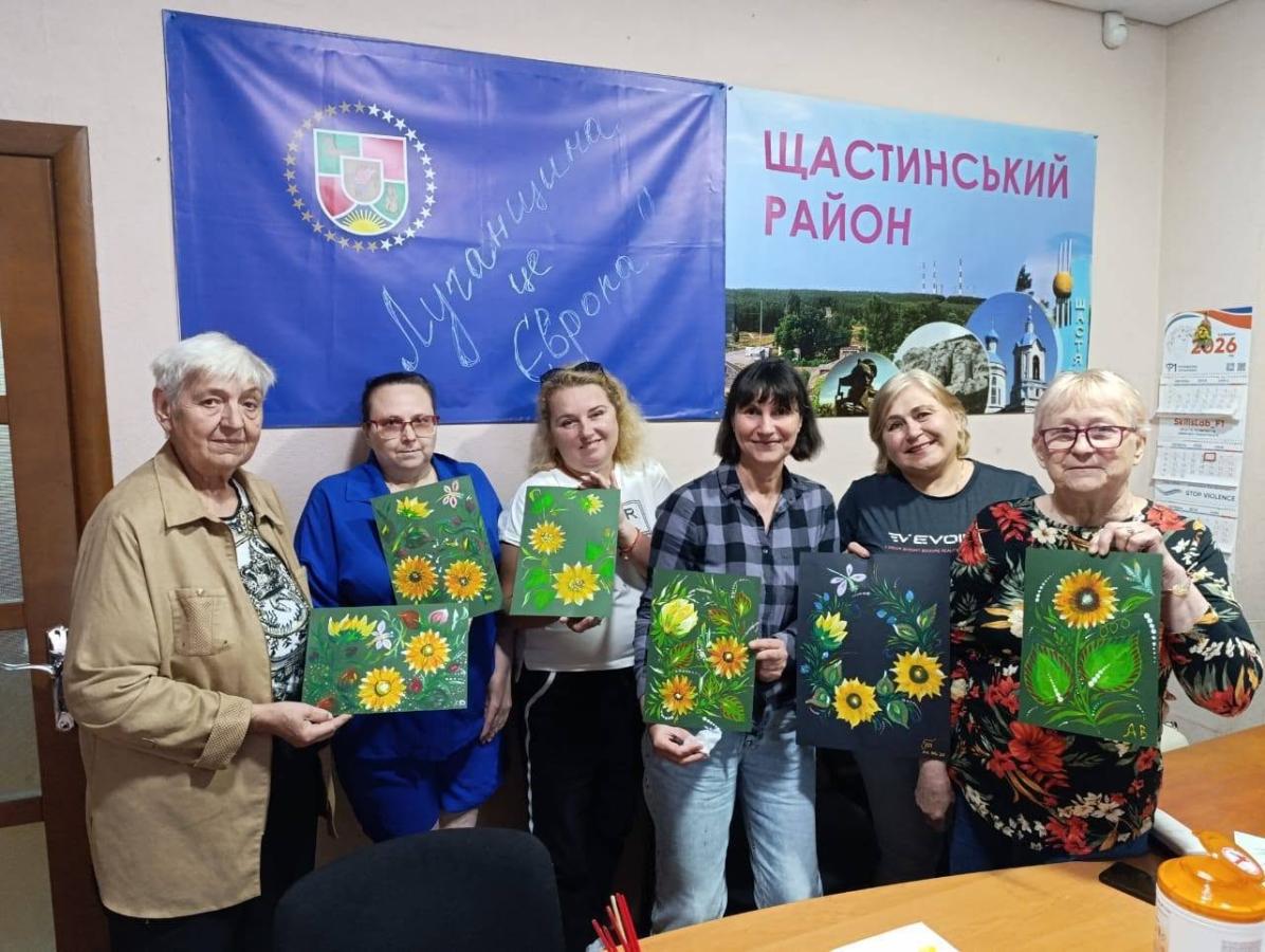 У приміщенні шість жінок різного віку стоять у ряд перед столом і тримають у руках свої малюнки з яскравими соняшниками на тлі прапора з написом «Луганщина це Європа» та плаката «Щастинський район»