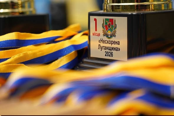 Спортивний кубок за перше місце з гербом Луганської області та написом «1 місце «Нескорена Луганщина» 2026» стоїть на столі поруч із купою розмитих на передньому плані синьо-жовтих нагородних стрічок.