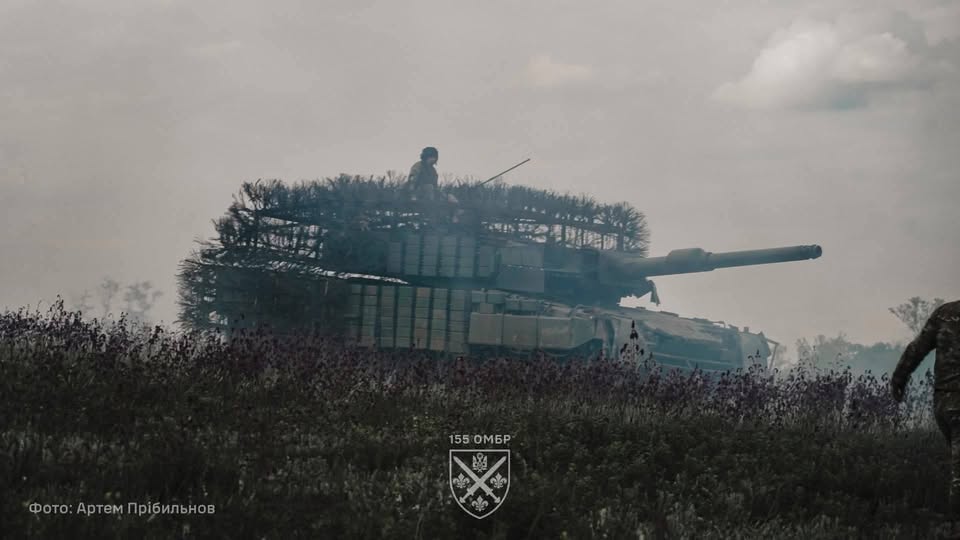 На фото зображено український танк Leopard 2 з масивним захисним навісом («мангалом») від дронів та блоками динамічного захисту, що стоїть у полі під хмарним небом