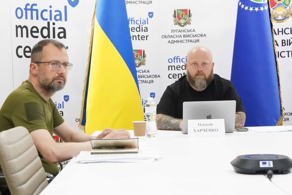 Двоє чоловіків сидять за білим столом під час засідання в офіційному медіацентрі Луганської обласної військової адміністрації на тлі прапорів України та відомства