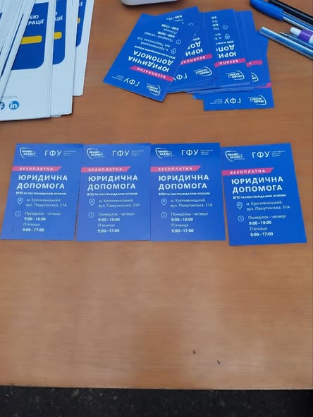 На дерев'яному столі розкладено сині інформаційні картки з текстом «БЕЗОПЛАТНА ЮРИДИЧНА ДОПОМОГА ВПО та постраждалим особам» від організацій «Право на захист» та «ГФУ», де зазначено адресу в місті Кропивницький та графік роботи.