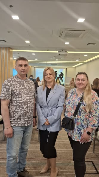 На фотографії троє людей — чоловік у футболка з візерунком та дві жінки у діловому й повсякденному вбранні — стоять поруч і посміхаються в конференц-залі.
