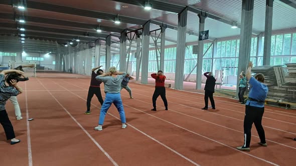 У великому критому легкоатлетичному манежі з біговими доріжками група людей старшого віку виконує фізичні вправи з розминки під керівництвом інструктора