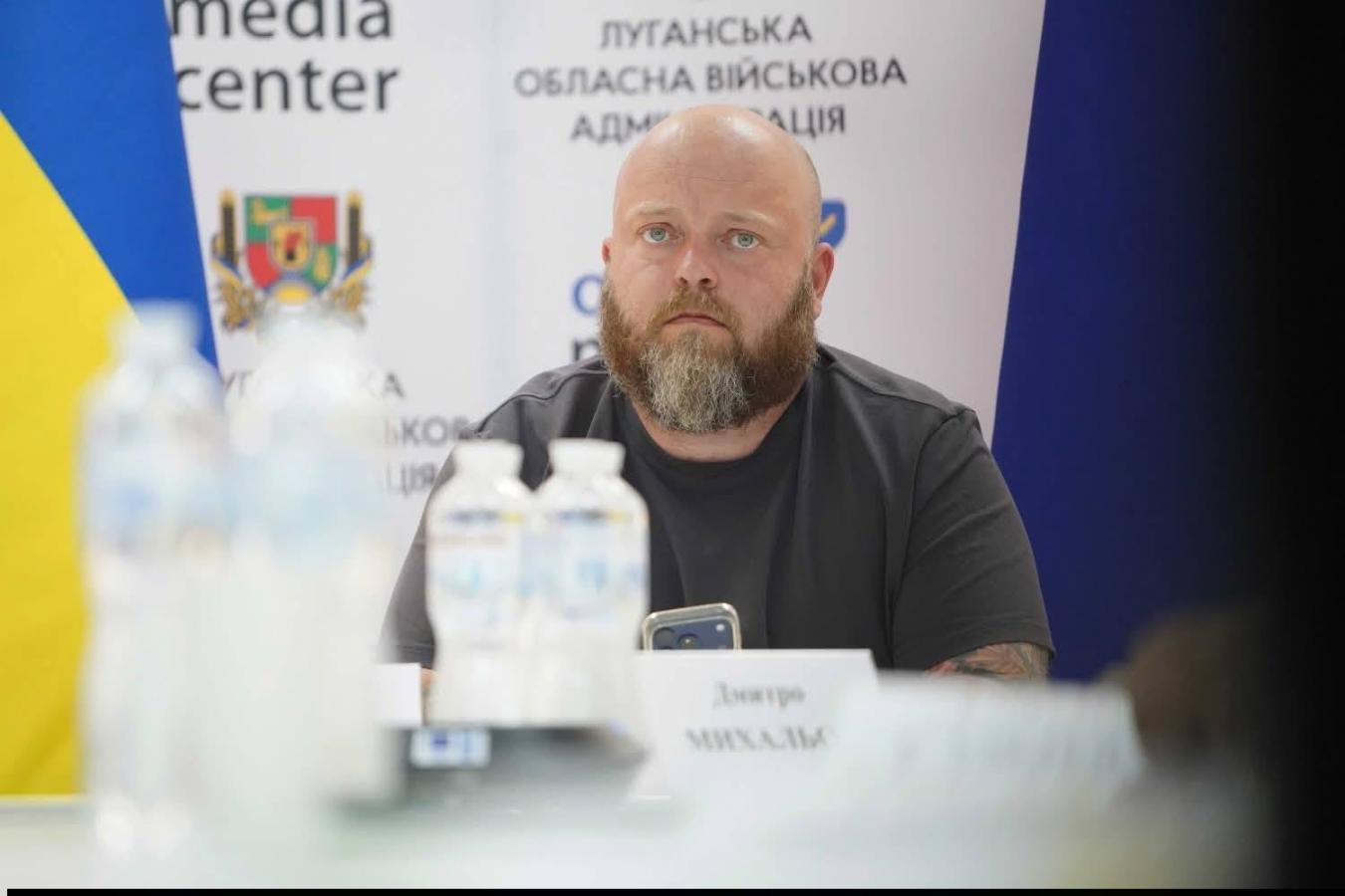 На тлі банера Луганської обласної військової адміністрації за столом із пляшками води сидить лисий чоловік із густою бородою у темній футболці, уважно дивлячись убік