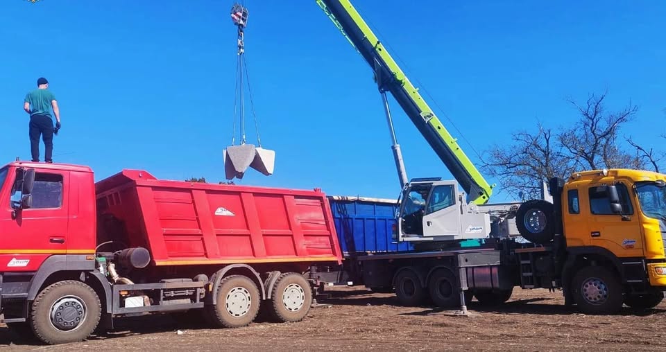 На тлі ясного синього неба автомобільний кран вантажить бетонні оборонні блоки («зуби дракона») у кузов великої червоної вантажівки, на даху кабіни якої стоїть чоловік