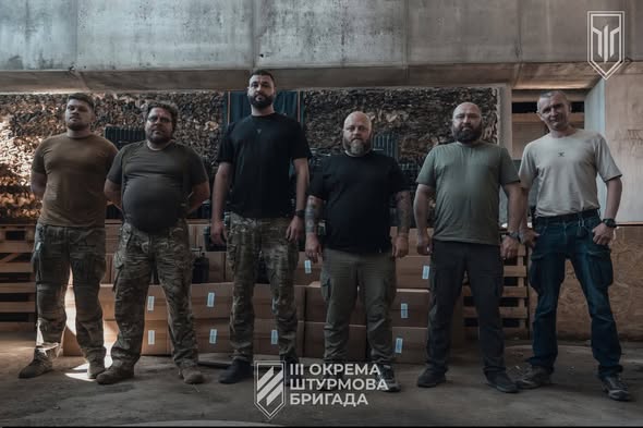На світлині у закритому приміщенні стоять у ряд шестеро чоловіків у військовому та повсякденному одязі на тлі картонних коробок і дров, а внизу та у верхньому правому куті розміщено емблему з написом «ІІІ ОКРЕМА ШТУРМОВА БРИГАДА».