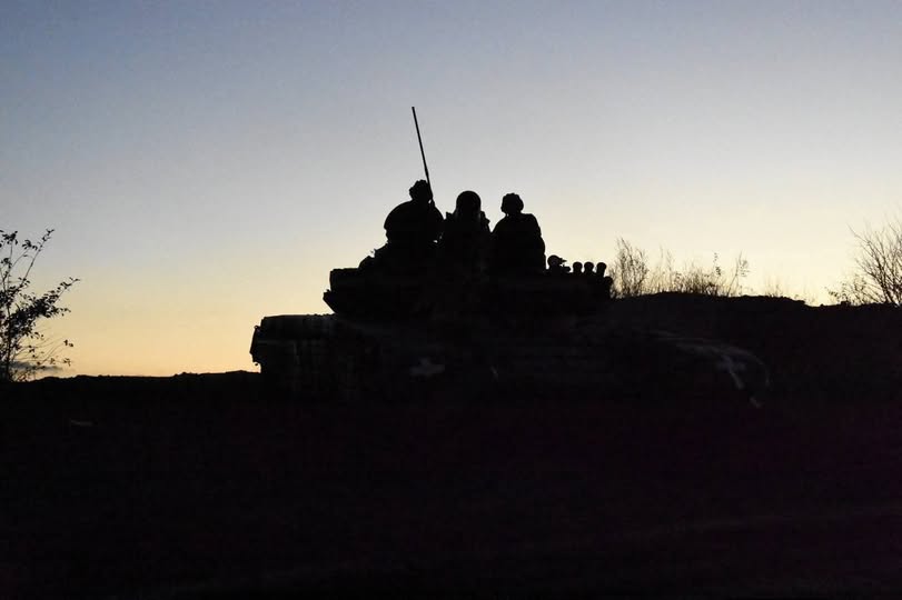 На зображенні на тлі вечірнього неба у світлі західу сонця видно темні силуети військового танка та кількох бійців, які сидять на його броні.