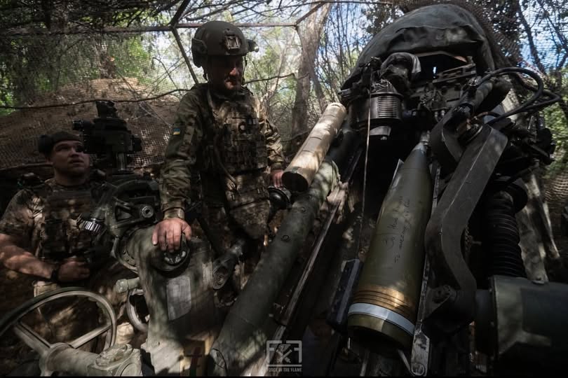 На зображенні під маскувальною сіткою в лісі двоє українських військових готують до пострілу артилерійську установку, великий снаряд якої завантажено в механізм.