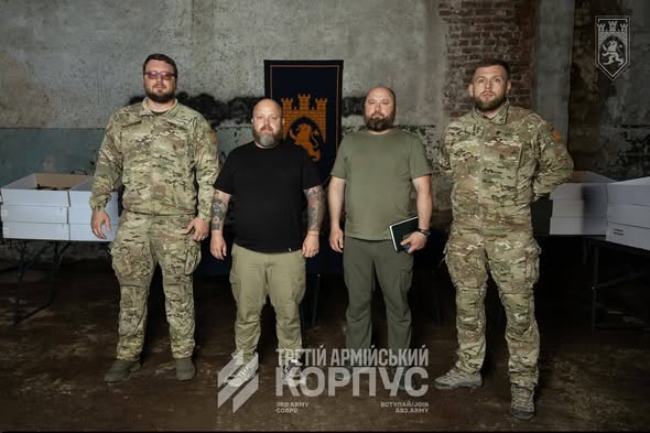 На фото зображено чотирьох чоловіків, двоє з яких у військовій формі камуфляжу «мультикам» та двоє у цивільному одязі мілітарі-стилю, які стоять у ряд у приміщенні на тлі прапора із зображенням лева та фортеці.