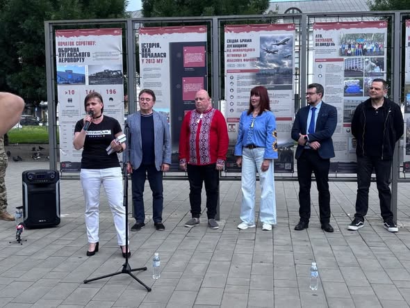 На зображенні по центру крупним планом показано українського військового у камуфляжній формі та коричневому береті, який тримає руку на зброї, на тлі інших бійців у строю, поверх яких по центру накладено великий жовтий напис «НАЦІОНАЛЬНИЙ СПРОТИВ»