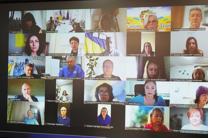 На фото зображено екран монітора з онлайн-нарадою у форматі відеоконференції, де у сітці з понад двадцяти вікон видно посадовців, медиків та представників громад Луганщини, деякі з яких мають підписи організацій та прапори на тлі.