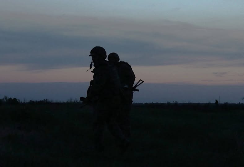 На фото зображено темні силуети двох військових у касках та зі спорядженням, які йдуть полем на тлі вечірнього або ранкового неба у сутінках.