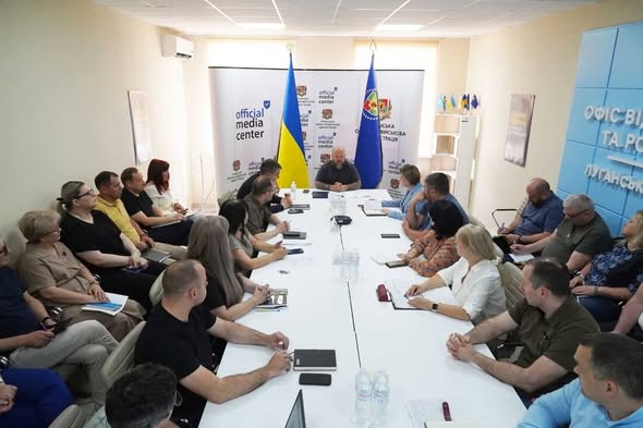 На фото зображено офіційне засідання або пресконференцію, де група людей сидить навколо довгого білого столу в кабінеті з українською символікою та прапорами
