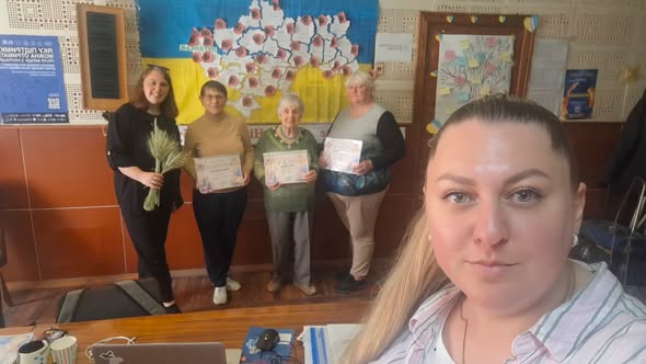 Група з п’яти жінок різного віку позує в кімнаті на тлі прапора та мапи України, при цьому троє з них тримають у руках грамоти, одна — пучок колосків, а жінка на передньому плані робить це селфі-фото.