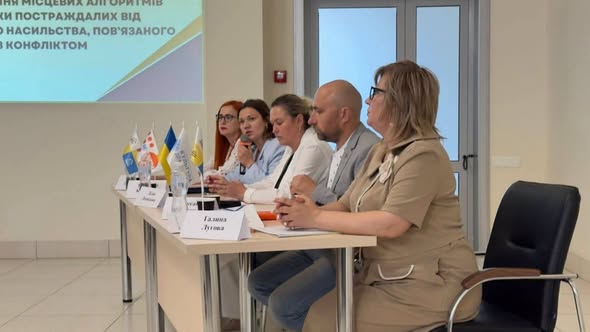 П'ятеро спікерів сидять за довгим столом із настільними прапорцями під час конференції, присвяченої допомозі постраждалим від насильства, пов'язаного з конфліктом.