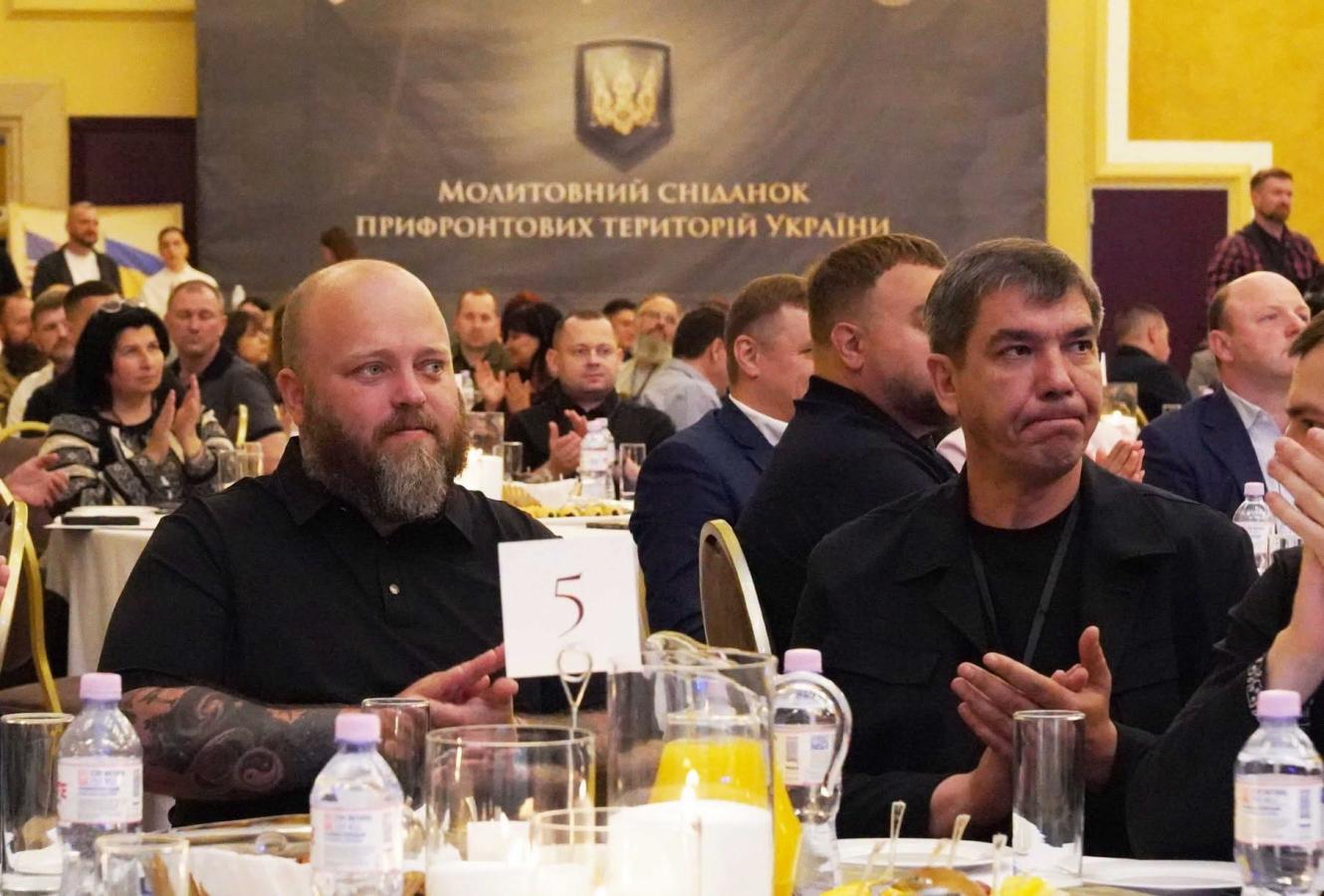 У великій залі на фоні банера з написом «Молитовний сніданок прифронтових територій України» за круглими сервірованими столами сидять численні учасники заходу, серед яких на передньому плані виділяються двоє чоловіків у чорному одязі, які аплодують.