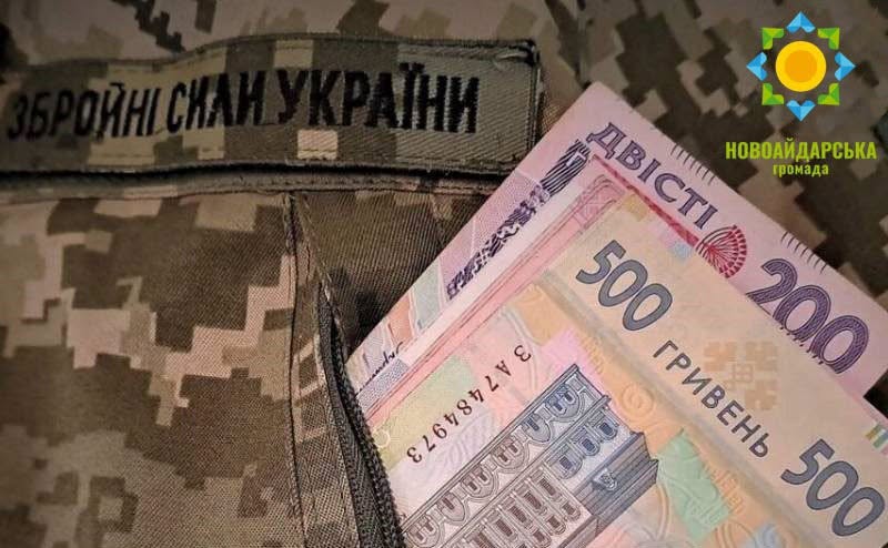 На зображенні показано банкноти номіналом 200 та 500 українських гривень, що виглядають з кишені військової камуфляжної форми з нашивкою «Збройні Сили України»
