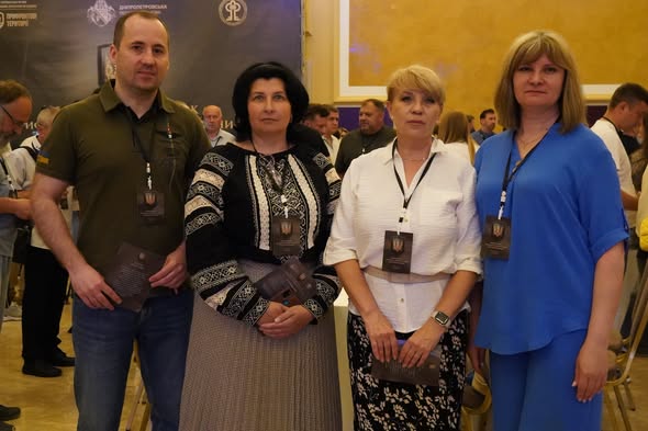 Один чоловік у зеленій футболці поло та три жінки в офіційно-діловому та традиційному одязі з бейджами на шиях стоять у ряд і позують для фото у великій залі на тлі інших людей та банера.