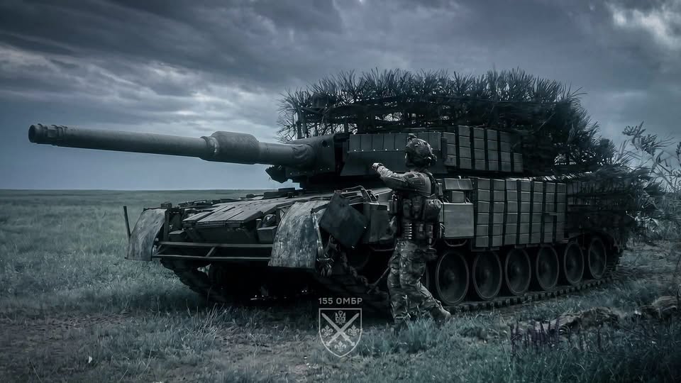 На фоні похмурого неба посеред зеленого поля стоїть замаскований гілками та блоками динамічного захисту військовий танк, біля якого спиною до глядача рухається солдат у повному бойовому спорядженні