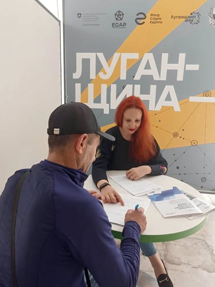 За круглим столом чоловік у кепці сидить спиною до камери та заповнює документи під наглядом жінки з яскравим рудим волоссям, а на задньому плані стоїть великий інформаційний банер із великим написом «ЛУГАНЩИНА»