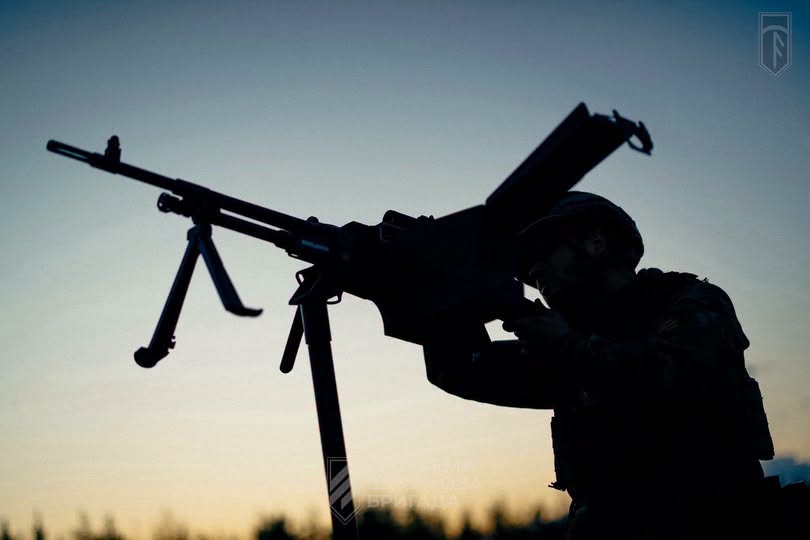 На тлі вечірнього неба знято темний силует українського військового у касці, який цілиться з великокаліберного кулемета, встановленого на станині-тринозі.