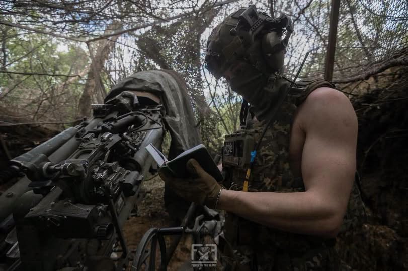 На зображенні військовослужбовець у касці, захисних навушниках та бронежилеті робить записи в блокноті, стоячи в замаскованому лісистому укритті поруч з артилерійською установкою.