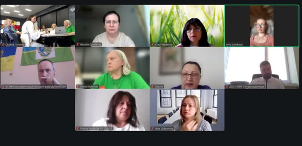 Скриншот екрана показує онлайн-зустріч у Zoom або схожій програмі, де у вікнах сітки розміщені обличчя десятьох учасників, переважно жінок, серед яких один чоловік сидить на тлі прапорів, а інша учасниця транслює відео з групової офлайн-зустрічі в офісі