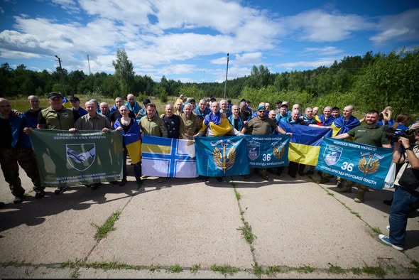 Велика група українських військовослужбовців та звільнених захисників позує на відкритій місцевості під синім небом, тримаючи в руках державні прапори України та різні військові стяги, зокрема 36-ї окремої бригади морської піхоти.