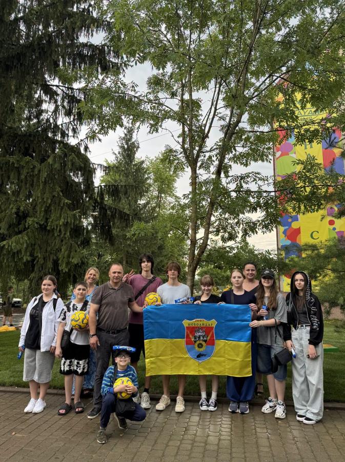 Група дорослих та дітей позує для спільного фото на вулиці біля зелених дерев, тримаючи синьо-жовтий прапор із гербом Новоайдара та кілька жовто-блакитних футбольних м'ячів.