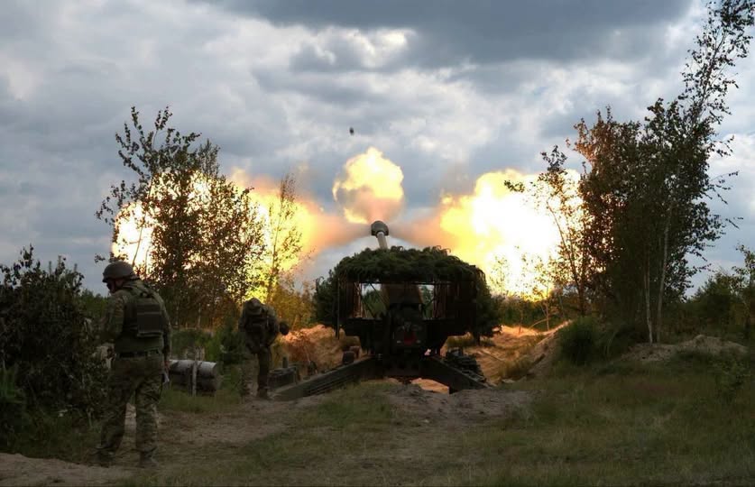 На зображенні зафіксовано момент потужного вогняного спалаху під час пострілу замаскованої артилерійської установки серед дерев, за яким зліва спостерігають двоє військових у камуфляжі