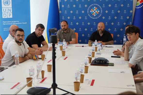 Група чоловіків сидить за великим білим Т-подібним столом під час офіційного засідання на тлі брендованих синіх банерів ПРООН та Конгресу місцевих та регіональних влад