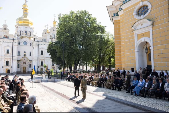 На світлині зображено офіційну церемонію на подвір'ї Києво-Печерської лаври, де Президент України Володимир Зеленський вручає державну нагороду військовому перед численними запрошеними гостями та військовослужбовцями на тлі величних білокам'яних храмів із золотими банями.