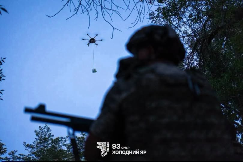 На світлині зображено силует українського військового зі зброєю на передньому плані, який спостерігає за великим гексакоптером у вечірньому небі, що переносить вантаж на тросі, із логотипом 93-ї ОМБр «Холодний Яр» у нижній частині кадру.
