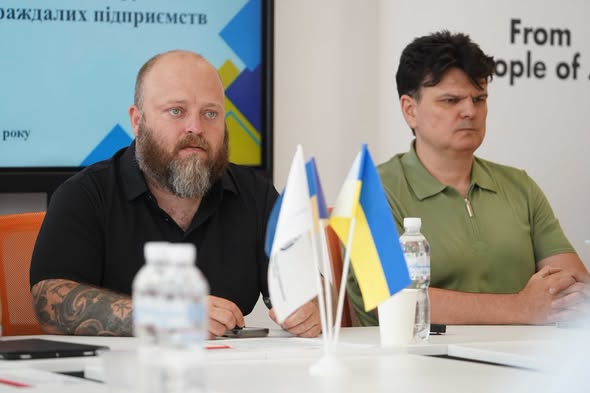 На зображенні двоє чоловіків сидять за столом під час офіційної зустрічі чи презентації, де ліворуч сидить бородатий чоловік із татуюваннями на руках у чорній сорочці, праворуч — чоловік у зеленій футболці-поло, а на передньому плані перед ними стоять маленькі прапорці України та інші настільні прапори поруч із пляшками води.